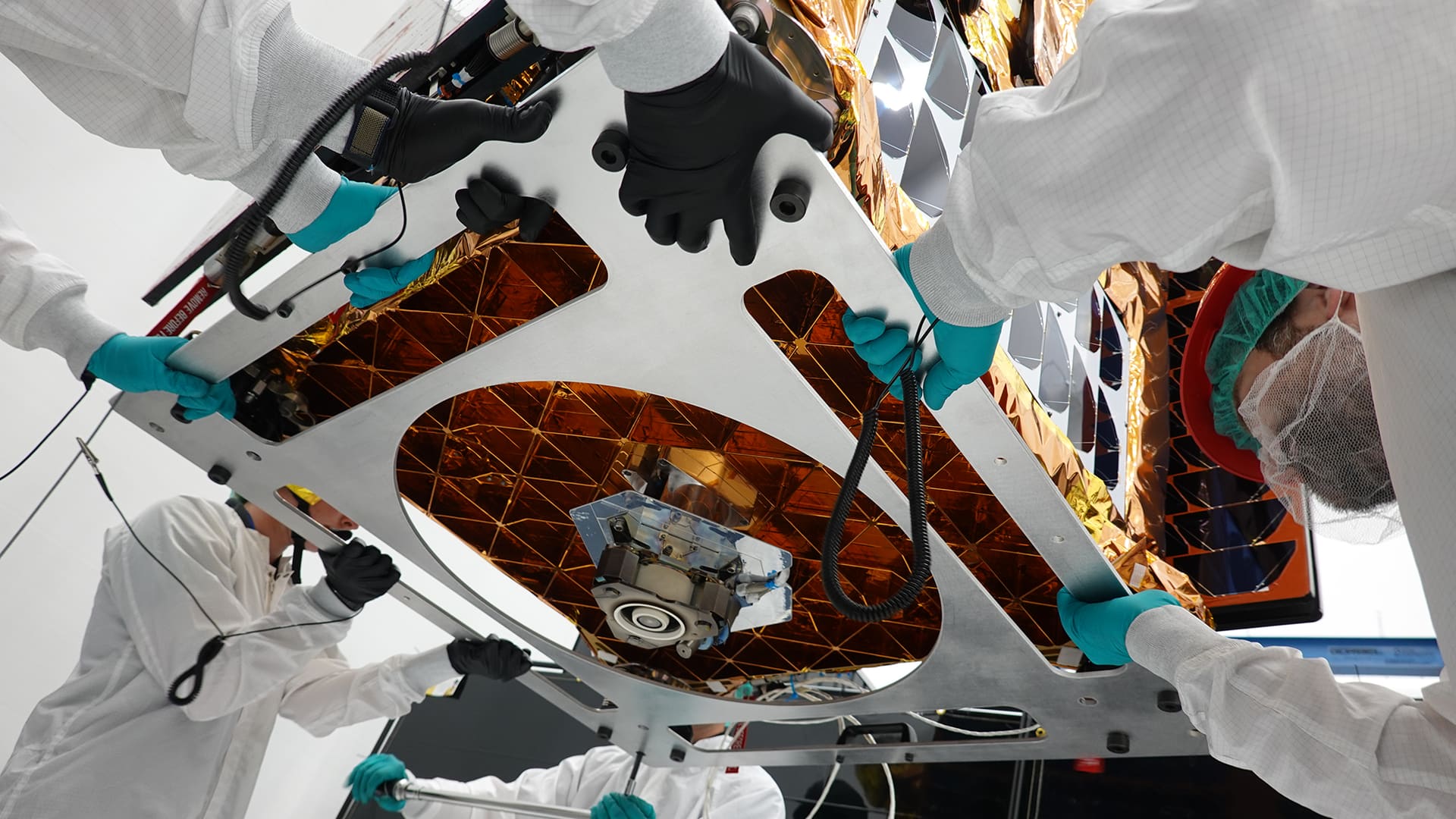 Four technicians in white suits and teal gloves handle the Otter spacecraft before launch.